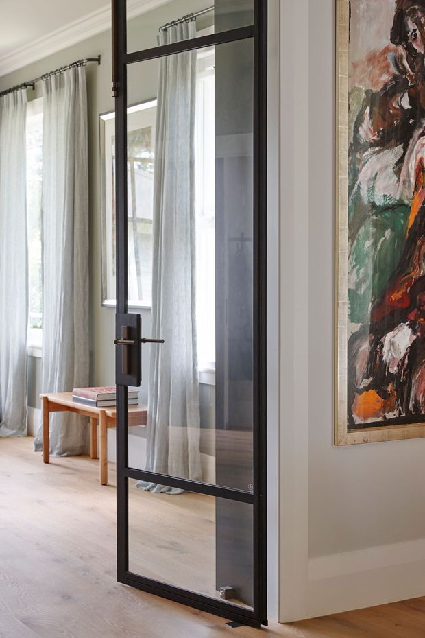 Interior view through a black-framed glazed steel-style pivot door into a bright room with tall sheer curtains, pale timber flooring, a small timber table, and a large abstract artwork on the adjacent wall. The space has white walls, traditional cornice detailing, and a refined contemporary-traditional feel.