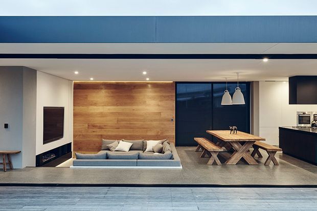 Open-plan contemporary living and dining area with a built-in timber feature wall, low grey sofa, black-framed glazing, exposed concrete-look flooring, pendant lights, and a compact white kitchen at right. A large opening connects the interior to an exterior deck or terrace in the foreground.