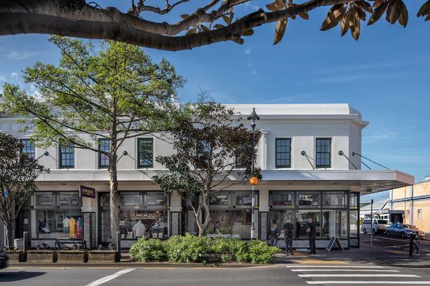 Street-facing view of a refined two-storey commercial building with a white rendered facade, classical horizontal cornice detailing, tall black-framed windows, and ground-floor retail shopfronts beneath a broad awning. Mature street trees, landscaped planting, pavement, and a pedestrian crossing are visible in the foreground.