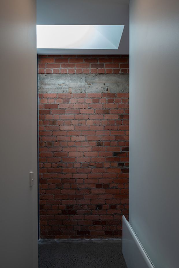 A narrow interior passage or small utility-like space with exposed red brick and a horizontal band of rough concrete forming the rear wall. Smooth light-grey side walls frame the space, with a small high-level frosted or translucent window providing daylight. The floor appears dark and textured, and a white radiator is visible along the lower right wall. The image has an industrial, moody character with strong contrast between the brickwork and painted surfaces.