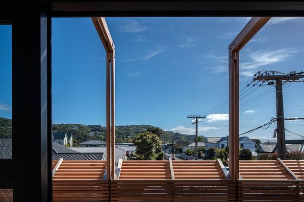 View from inside through large black-framed glazing toward a timber outdoor terrace or balcony with horizontal slatted screening and substantial timber posts. Beyond are pitched-roof houses, utility poles, and a wooded hillside under a clear blue sky. The image is dominated by the framed view and exterior deck structure.
