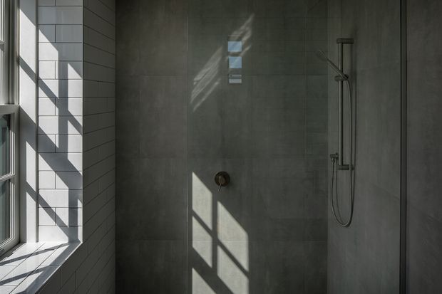 A minimalist bathroom shower area with large dark grey wall tiles, a frameless glass shower enclosure, and a slim dark metal shower rail and controls. White rectangular wall tiles surround a tall window on the left, casting strong geometric sunlight and shadows across the tiled surfaces.