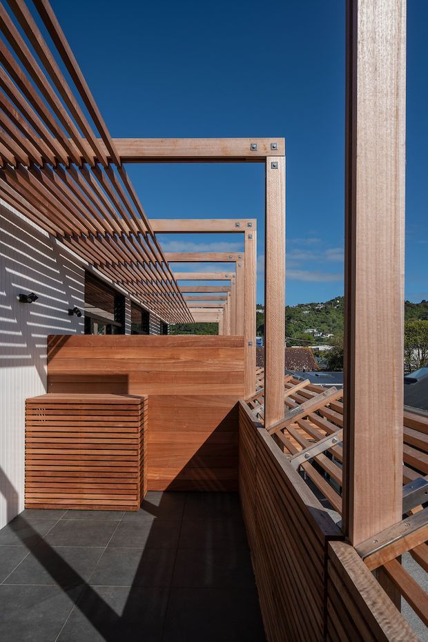 A sunlit timber-lined balcony or terrace with strong contemporary detailing. Repeated exposed timber pergola beams and vertical posts frame the space, casting striped shadows across white cladding and dark tiled flooring. Timber privacy screens and balustrades create a warm, structured outdoor room, with suburban rooftops and greenery visible beyond.