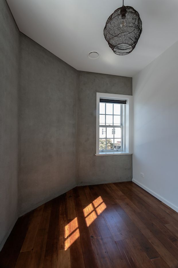 A narrow, unfurnished interior room with dark timber floorboards, a textured grey cement-like feature wall, and white painted walls and ceiling. A small multi-pane window provides daylight, casting a strong geometric reflection across the floor. A black woven pendant light hangs from the ceiling, and a recessed circular downlight is also visible. The room has a simple, restrained contemporary character.
