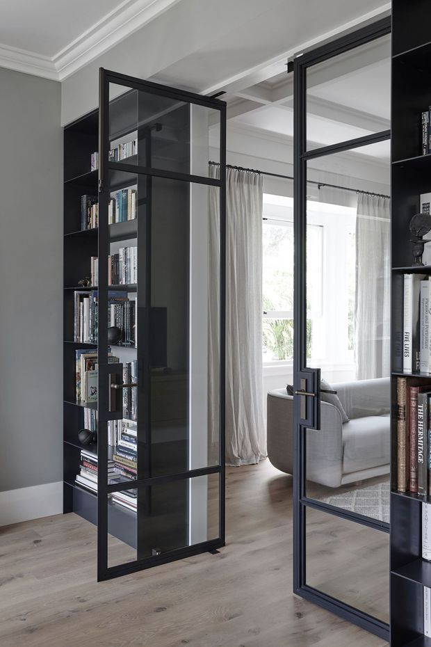 Interior view through black-framed glazed steel doors, with integrated dark bookcases on both sides. The doors open from a study or library into a bright living room featuring a pale upholstered sofa, light curtains, large windows, and light timber flooring. White decorative ceiling mouldings and grey painted walls add a refined traditional character, while the slim dark metal glazing creates a contemporary contrast.