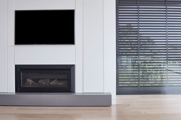 Modern living room media wall featuring a wall-mounted black television above a built-in linear fireplace with dark surround and grey hearth. Full-height white built-in wall panels frame the fireplace, while large glazing with dark horizontal blinds occupies the right side. Pale timber flooring and restrained neutral finishes create a clean, contemporary interior.