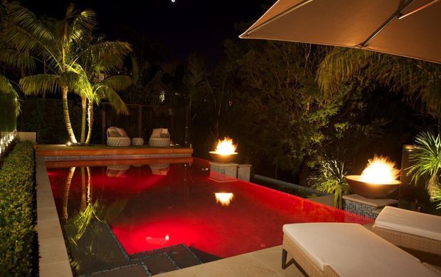 Night-time view of a luxurious outdoor pool area with vivid red underwater lighting, dark paving, tropical planting, sculptural fire bowls, sun loungers, and a large shade canopy. The pool and surrounding entertaining space create a dramatic resort-like atmosphere.