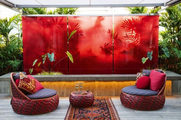 A vibrant semi-enclosed outdoor lounge with two rounded woven armchairs, patterned cushions, a small round table, and a decorative rug. A raised planter with tropical greenery sits behind a striking translucent red screen, beneath a glazed or louvred roof structure.