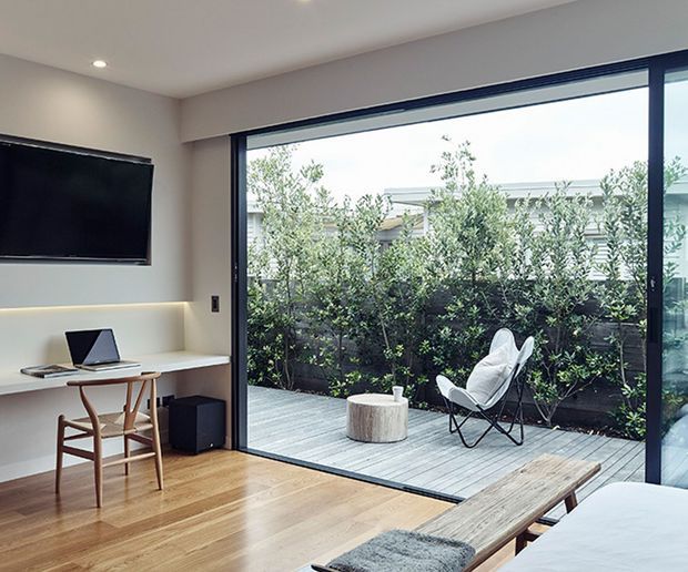 A bright, compact contemporary living and work area opening onto a timber deck through expansive black-framed sliding glass doors. The interior has pale walls, recessed ceiling lights, warm timber flooring, a wall-mounted television, a simple desk with laptop, and a low bench or table. Outside, a small deck is furnished with a white sling chair and cylindrical timber coffee table, with dense screening planting beyond.