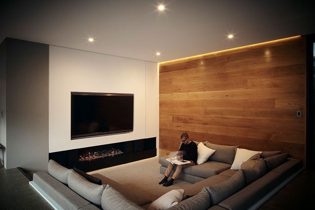 A contemporary living room centered on a large U-shaped sectional sofa, with a wall-mounted television and long linear fireplace set into a dark surround. A full-height warm timber feature wall with concealed cove lighting contrasts with smooth light walls and a dark floor. Recessed ceiling lights provide focused illumination, creating an intimate media-oriented lounge.