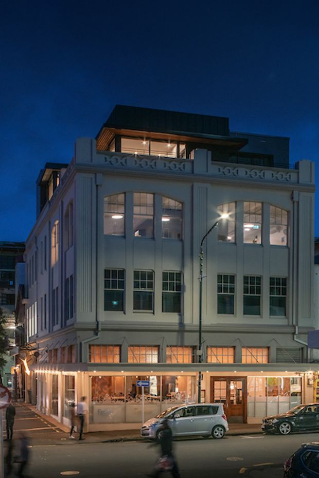 Twilight street-level view of a multi-storey historic commercial building with a pale ornate facade, tall multi-pane windows, decorative pilasters and parapet detailing. The ground floor contains a brightly lit glazed shopfront or hospitality space, while a contemporary rooftop addition with dark cladding and an illuminated terrace sits above the original structure. Urban street activity and parked vehicles are visible in the foreground.