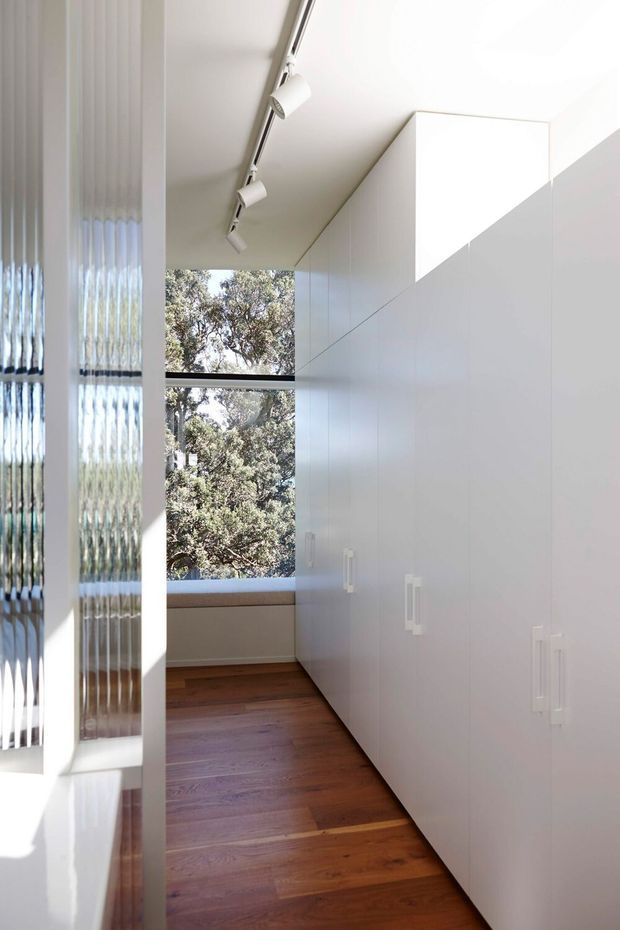 A narrow, bright built-in storage corridor or walk-in wardrobe with full-height glossy white cabinetry and simple white handles along the right wall. Warm timber flooring runs toward a large window overlooking trees, while a textured glass partition and ceiling-mounted track spotlights add architectural detail.