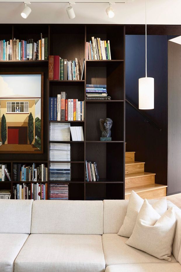 A refined living room featuring a large dark timber built-in bookshelf filled with books and magazines, a framed architectural artwork, a pale upholstered sectional sofa with cushions, and a concealed staircase with light timber treads. Track lighting and a suspended pendant provide illumination, while a dark blue wall adds contrast.