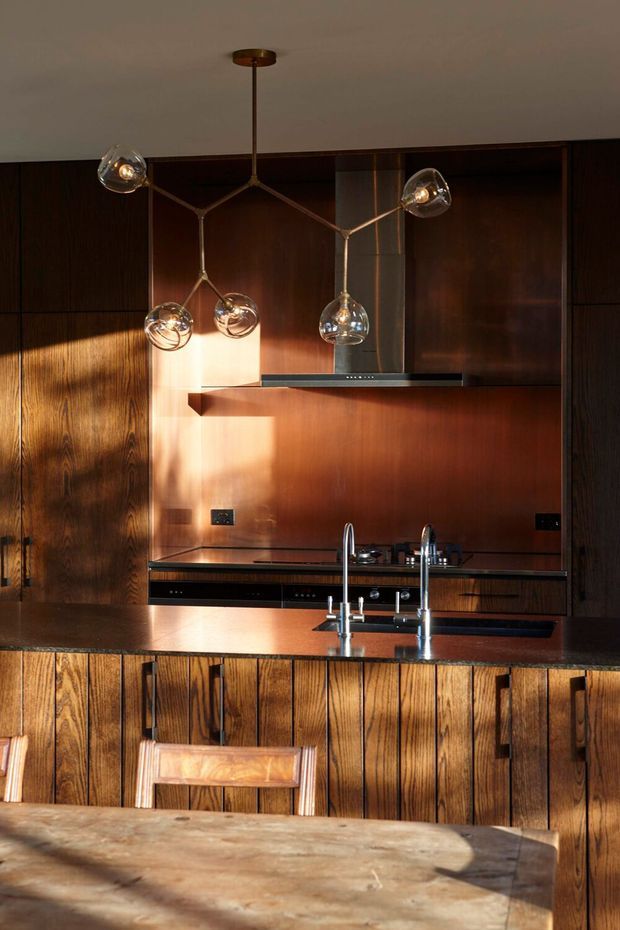 Warm, moody kitchen interior with extensive dark timber wall panelling and fluted timber island cabinetry. A dark stone benchtop, copper-toned splashback, integrated cooktop and rangehood, undermount sink with twin faucets, and sculptural multi-globe pendant lighting define the space. Strong sunlight creates dramatic highlights across the timber surfaces.