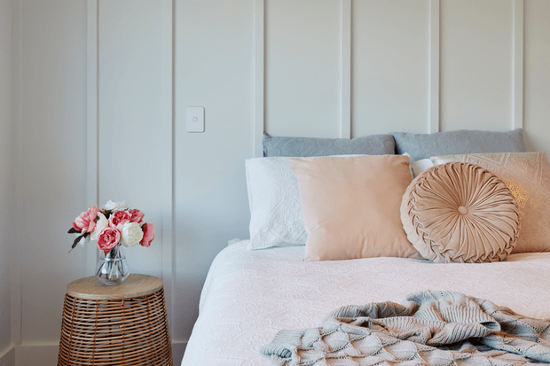 A softly styled bedroom corner featuring a neatly made bed with layered white, pale peach, and blush bedding, multiple decorative pillows, and a textured throw. The bed is set against light grey-blue vertical wall panelling, with a small woven rattan side table holding a glass vase of flowers.