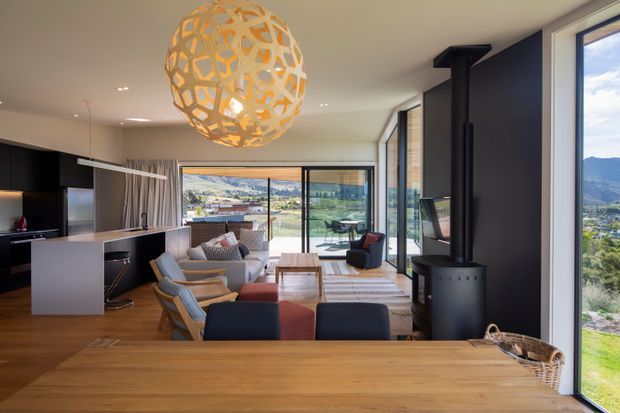 Open-plan contemporary living and dining area with a modern kitchen, timber dining table, lounge seating, large sliding glass doors, and expansive mountain views. A freestanding black wood-burning stove and sculptural pendant light provide focal points, while warm timber flooring and furniture create a welcoming interior.