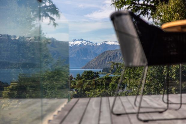 A timber outdoor deck viewed through or beside glazing, with a blurred outdoor chair in the foreground and a lake, rugged mountains, and snow-capped peaks beyond. The scene is bright and natural, suggesting a mountain or lakeside terrace.