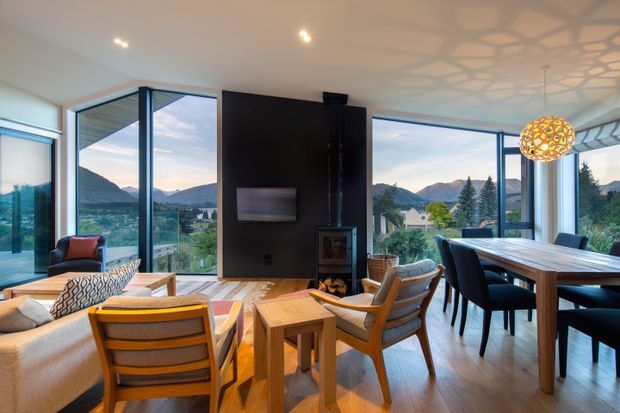Bright, open-plan living and dining area with extensive floor-to-ceiling glazing overlooking a mountainous landscape. The room features warm timber flooring and furniture, a black feature fireplace wall with integrated television, contemporary seating, and a sculptural pendant light casting patterned shadows across the ceiling.