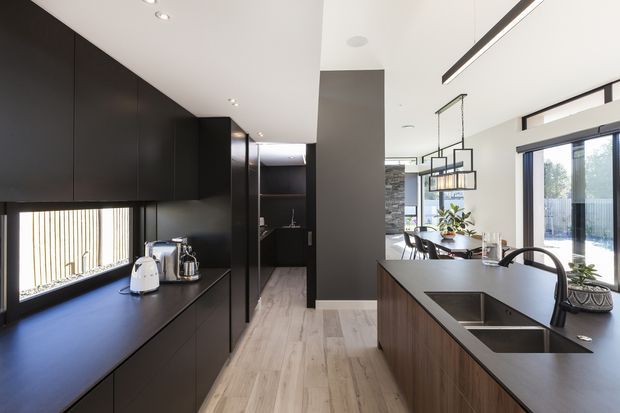 A contemporary open-plan kitchen with extensive matte black cabinetry, dark benchtops, a timber island with an integrated double sink, and pale timber flooring. The kitchen opens to a dining and living area with large glazed doors, black-framed windows, pendant lighting, and abundant natural light.