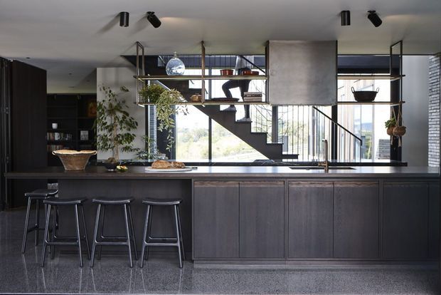 A sophisticated open-plan kitchen dominated by a long dark built-in island with a stone benchtop and three black bar stools. Open timber-and-metal shelving separates the kitchen from a stairway, with visible plants, ceramics and decorative objects. The room has dark cabinetry, a polished grey floor, pale ceiling, track lighting and expansive glazing around the stairwell, creating a refined, contemporary entertaining space.
