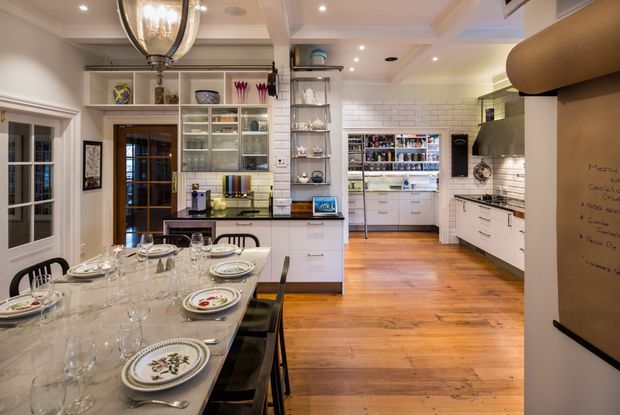A spacious traditional-style kitchen and dining area with warm timber flooring, white shaker-style cabinetry, open glass-front shelving, white subway-tile splashbacks, and extensive built-in storage. A long dining table is set in the foreground beneath a decorative pendant light, while a pantry or utility storage area is visible at the rear. The room has layered lighting, detailed trim, and a warm, refined feel.