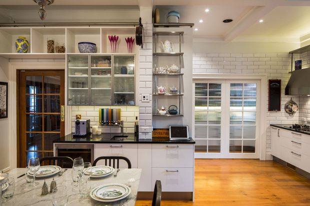 A bright, traditional-style kitchen with white cabinetry, glass-front display cupboards, open shelving, and white subway-tile walls. The room includes a dark stone island or peninsula, integrated appliances, a gas cooktop with rangehood, timber flooring, and French doors opening to the outside. A dining table is set in the foreground.