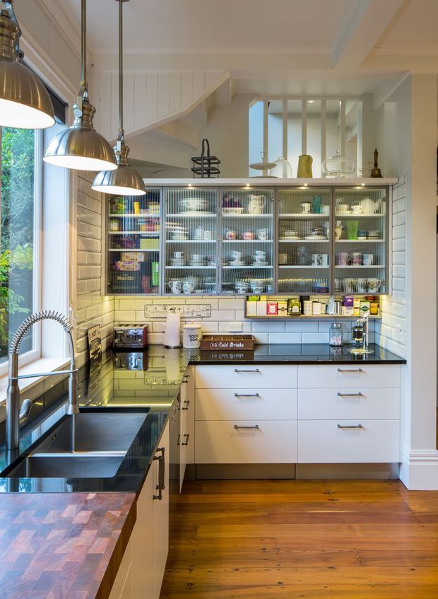 A bright, compact traditional kitchen with white vertical wall panelling, white subway-tile splashback, extensive white cabinetry, glass-fronted upper storage, dark stone-look benchtops, and warm timber flooring. Three large metal pendant lights hang over the island, while a deep undermount sink sits beside a tall window. The kitchen includes open shelving and display cabinetry for crockery and pantry items, with polished chrome hardware and a warm, practical farmhouse-inspired feel.