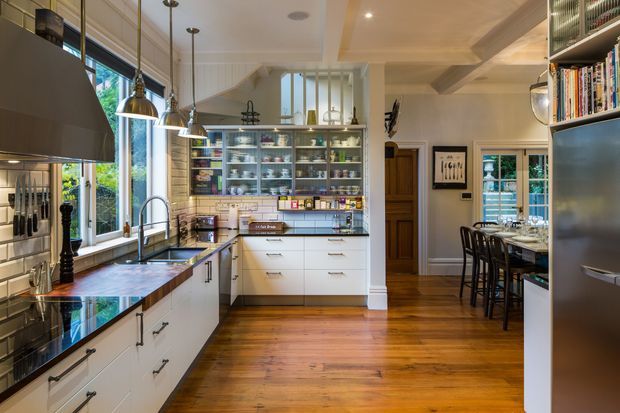 A warm, spacious traditional kitchen with white built-in cabinetry, a dark timber island benchtop, stainless-steel appliances, pendant lighting, open display shelving, and extensive windows. A dining area is visible beyond the kitchen, with timber flooring throughout and a coffered ceiling adding architectural detail.