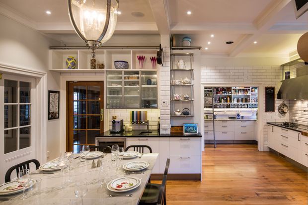 A spacious, traditional-style kitchen and dining area with warm timber flooring, white shaker-style cabinetry, glass-front display cupboards, and white subway-tile splashbacks. A large marble-look dining table is set with plates and glassware beneath an ornate pendant light. The kitchen includes extensive storage, open shelving, integrated appliances, and a walk-in pantry visible at the rear.