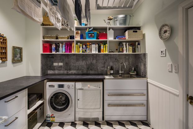 A compact, highly organised laundry room with white built-in cabinetry, dark stone-look benchtops and backsplash, open shelving filled with cleaning supplies and baskets, a front-loading washing machine and dryer, a utility sink, and hanging laundry overhead. The room features decorative wall panelling, a wall clock, and a bold black-and-white geometric tiled floor.