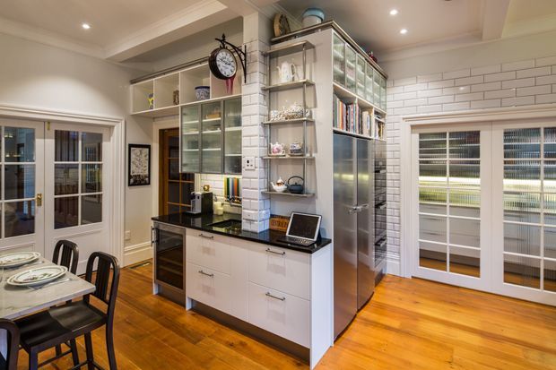 Bright, compact open-plan kitchen and dining area with white painted brick tile walls, white built-in cabinetry, black stone-look benchtops, glass-front display shelving, stainless steel appliances, and warm timber flooring. A dining table sits to the left, while glazed doors and plantation-style shutters provide architectural detail and natural light.