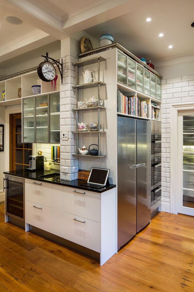 A compact, highly detailed kitchen with white subway-tile walls, glossy white lower cabinetry, black benchtops, stainless-steel appliances, and timber flooring. A tall integrated storage and refrigerator wall includes ribbed glass-front cabinets and open shelving displaying books and ceramics. Decorative shelving, a wall clock, recessed lighting, and a glass-panel door complete the warm, eclectic fit-out.