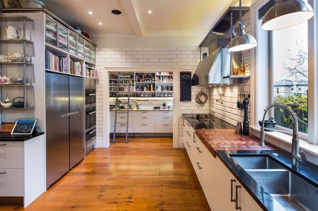 A bright, well-appointed galley kitchen with warm timber flooring, white subway-tile walls, extensive white cabinetry, open shelving, a large stainless-steel refrigerator, dark stone benchtops, a timber butcher-block preparation area, and a double-bowl sink beneath a large window. A walk-in pantry or scullery is visible at the rear, with pendant lighting and integrated storage throughout.
