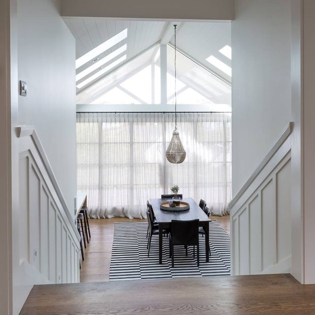 Bright dining room viewed from a stair landing, with a dark rectangular dining table and chairs on a black-and-white striped rug. Floor-to-ceiling sheer curtains, a woven pendant light, white geometric stair wall panelling, and a dramatic glazed pitched ceiling with skylights define the space.
