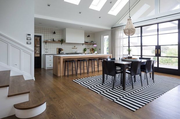 Bright open-plan kitchen and dining area with a large timber island, white tiled splashback, white cabinetry, skylights, black-framed glazed doors, a timber dining table with black chairs, and a bold black-and-white striped rug. A curved timber staircase and pendant lighting add architectural and decorative interest.