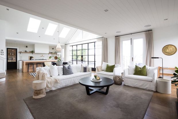 Bright open-plan living room with a large white slipcovered sofa and matching armchairs arranged around a black round coffee table, opening directly to a tiled kitchen with a timber island. The room features a white-lined pitched ceiling with skylights, expansive black-framed glazing, pale curtains, timber flooring, and a large neutral rug. The palette is light and airy with muted green accents.