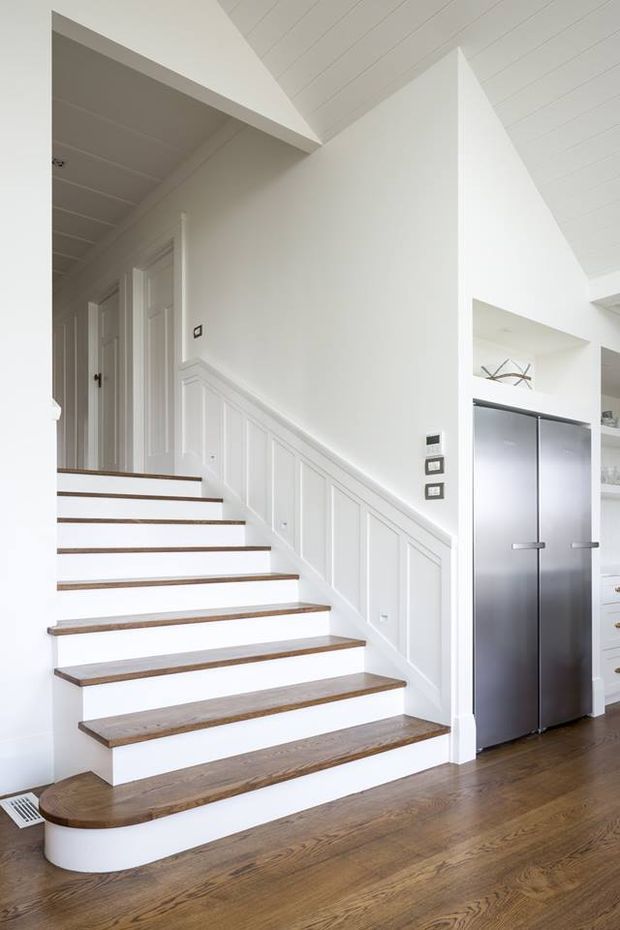 Bright white interior featuring a staircase with natural timber treads, white painted risers, and decorative white wall panelling beneath the stair. The stair rises beside a hallway with several white paneled doors and opens toward a kitchen area with a large dark refrigerator and white cabinetry. Wide-plank timber flooring and a white paneled ceiling create a clean, refined transitional look.