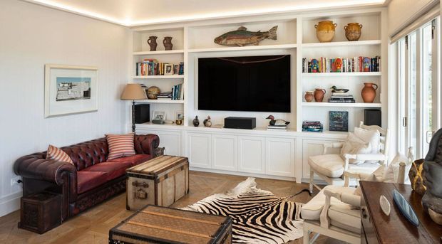 Warm, traditionally styled living room with built-in white entertainment cabinetry and open shelving surrounding a wall-mounted television. The room includes a deep red leather sofa, cream upholstered armchairs, a large timber coffee table, decorative ceramics and books, and wide windows with blinds. Recessed cove lighting and timber flooring create a refined, comfortable atmosphere.