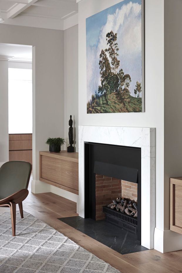 A refined living room centered on a traditional fireplace with a veined white marble surround, black firebox, and exposed brick interior. The room has pale grey walls, white architectural trim and ceiling beams, warm timber flooring, built-in timber cabinetry, a mid-century style leather lounge chair, patterned rug, and a large landscape painting above the mantel.