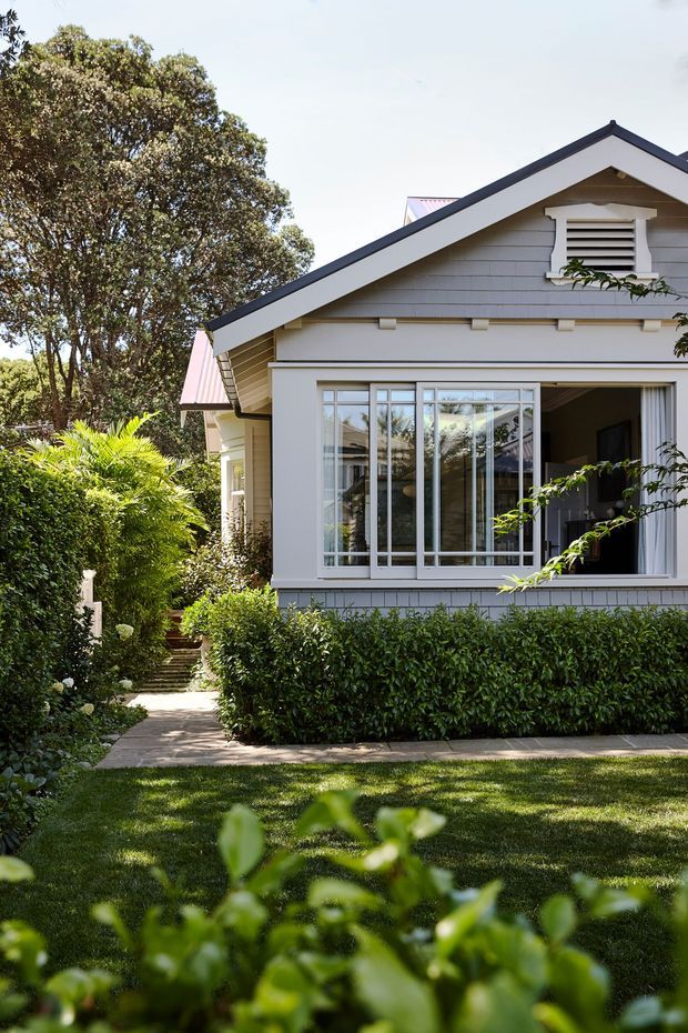 Exterior view of a light-painted, gable-roofed residential house with horizontal weatherboard cladding, dark roof edging, decorative gable vent, and large glazed windows. A manicured garden with hedges, palms, mature trees, lawn, and a paved path surrounds the facade. The open window/door reveals a small portion of the interior.