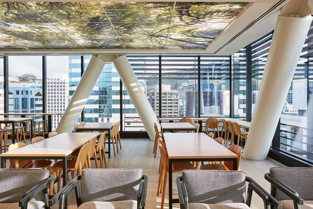 Bright contemporary commercial dining or lounge interior on an elevated urban level, with extensive floor-to-ceiling glazing, city views, exposed angled structural columns, timber tables and chairs, and upholstered armchairs. A dramatic backlit forest-canopy ceiling graphic adds a natural visual feature, while horizontal external sun-shading elements are visible across the glazing.