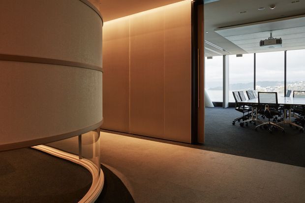 A refined modern commercial office interior viewed from a softly lit reception or circulation area. A curved illuminated architectural feature with layered cylindrical forms occupies the foreground, while a glazed boardroom with a long conference table, office chairs, ceiling-mounted projector and expansive city-facing windows is visible beyond. Warm timber wall panelling, concealed lighting and dark carpet create a sophisticated, understated atmosphere.