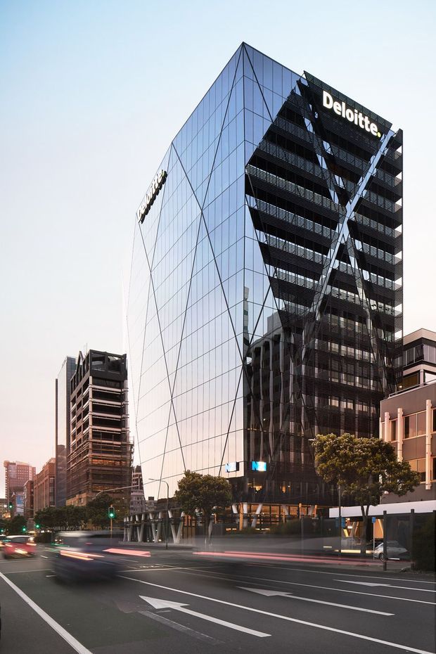 Street-level architectural photograph of a tall contemporary commercial office building with a sharply angled glass curtain-wall facade, dark external louvres and projecting structural elements, prominently displaying Deloitte signage. The building stands within a dense urban streetscape alongside other office buildings, trees, traffic lanes and illuminated street signals, photographed at dusk with motion-blurred vehicles.