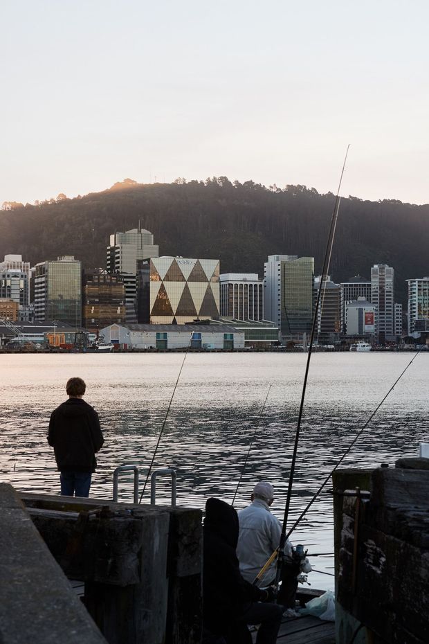 A waterfront urban scene viewed from a timber-and-concrete pier or dock, with people fishing in the foreground and a calm harbor across the water. The opposite shore has a dense cluster of contemporary mid- and high-rise commercial buildings backed by a wooded hillside. The image has soft, muted evening light and a documentary architectural-photography feel.