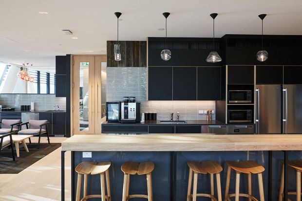 A contemporary open-plan kitchen dominated by a long island bench with a light timber top and dark integrated cabinetry. The island incorporates multiple timber bar stools and faces a full-height run of black cabinetry with integrated ovens, microwave, and stainless-steel refrigerator. A tiled splashback, sink, pendant lights, timber-framed glazing, and an adjacent dining/lounge area are visible.