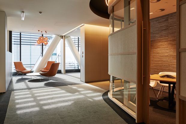 A contemporary commercial interior showing an open lounge and meeting area. The space has grey carpet, warm timber wall and door finishes, curved glazed partitions with upholstered acoustic panels, orange leather lounge chairs, and a small meeting room visible through the glass. Strong daylight enters through expansive windows, with exposed diagonal structural braces and decorative pendant lighting creating a refined office atmosphere.