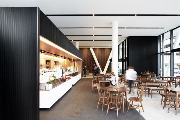 A bright contemporary hospitality interior, likely a café or restaurant, with a long illuminated service counter and open shelving on the left, numerous timber tables and spindle-back chairs, dark ribbed wall panels, white structural columns, exposed dark timber feature walls, and expansive black-framed windows. Linear recessed ceiling lights and a polished concrete floor create a clean, modern commercial atmosphere.