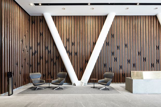 A refined contemporary reception or lobby space with dramatic full-height dark timber slat walls featuring repeating geometric inlays. Large white structural V-shaped columns create a bold focal point, while low modern lounge chairs and small round tables form seating areas around a light stone or concrete reception desk. A pale polished floor and recessed ceiling lighting contribute to the sophisticated commercial interior.