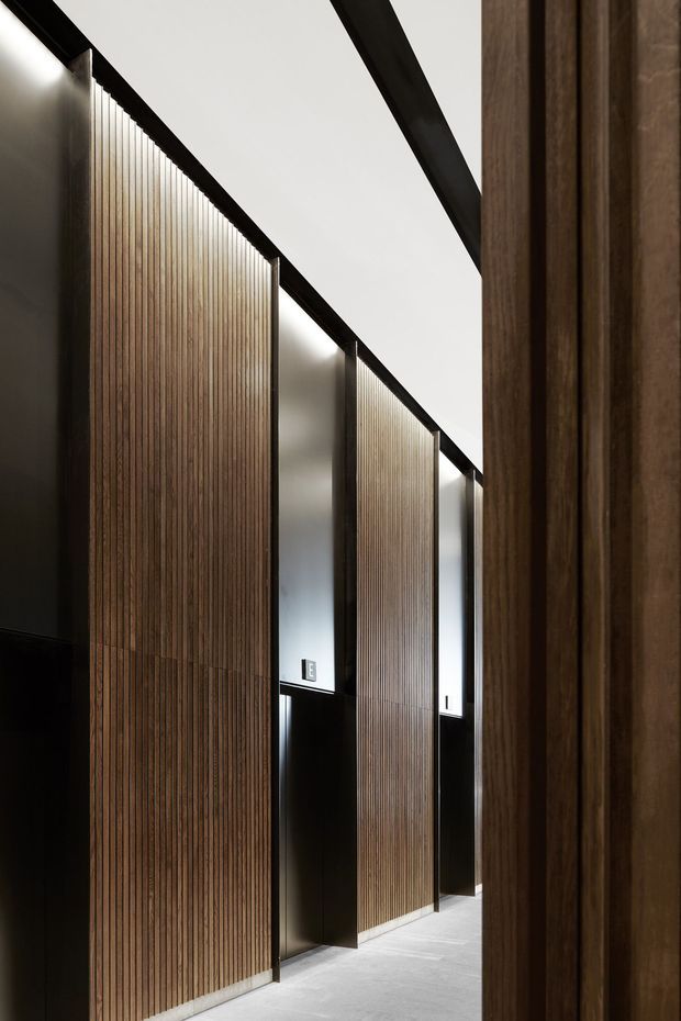 A narrow, highly refined interior corridor lined with tall vertical timber slat wall panels and dark metal framing. Recessed linear lighting creates a warm glow along the panel edges, while pale smooth flooring and a white ceiling provide contrast. The composition emphasizes repetition, strong vertical lines, concealed doors or joinery, and minimalist detailing.