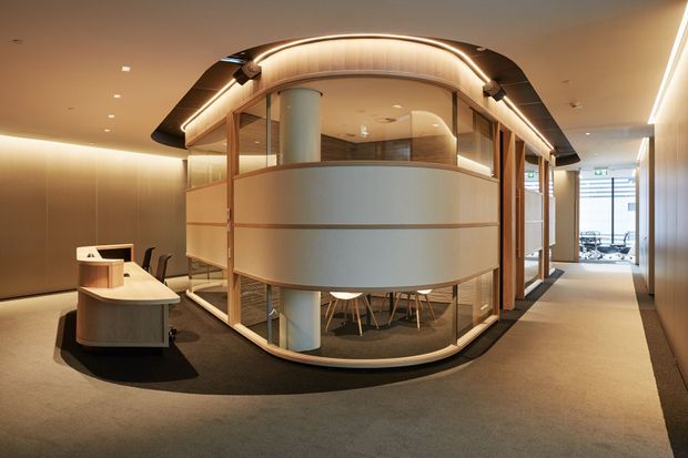 A refined contemporary commercial interior featuring a curved, glass-enclosed meeting or workspace pod with warm timber framing, frosted privacy panels, integrated ambient LED lighting, and cylindrical structural columns. A curved reception desk sits to the left, with carpeted circulation space and additional glazed work areas beyond. The palette is warm beige, taupe, timber, and soft grey, creating a polished and welcoming office environment.